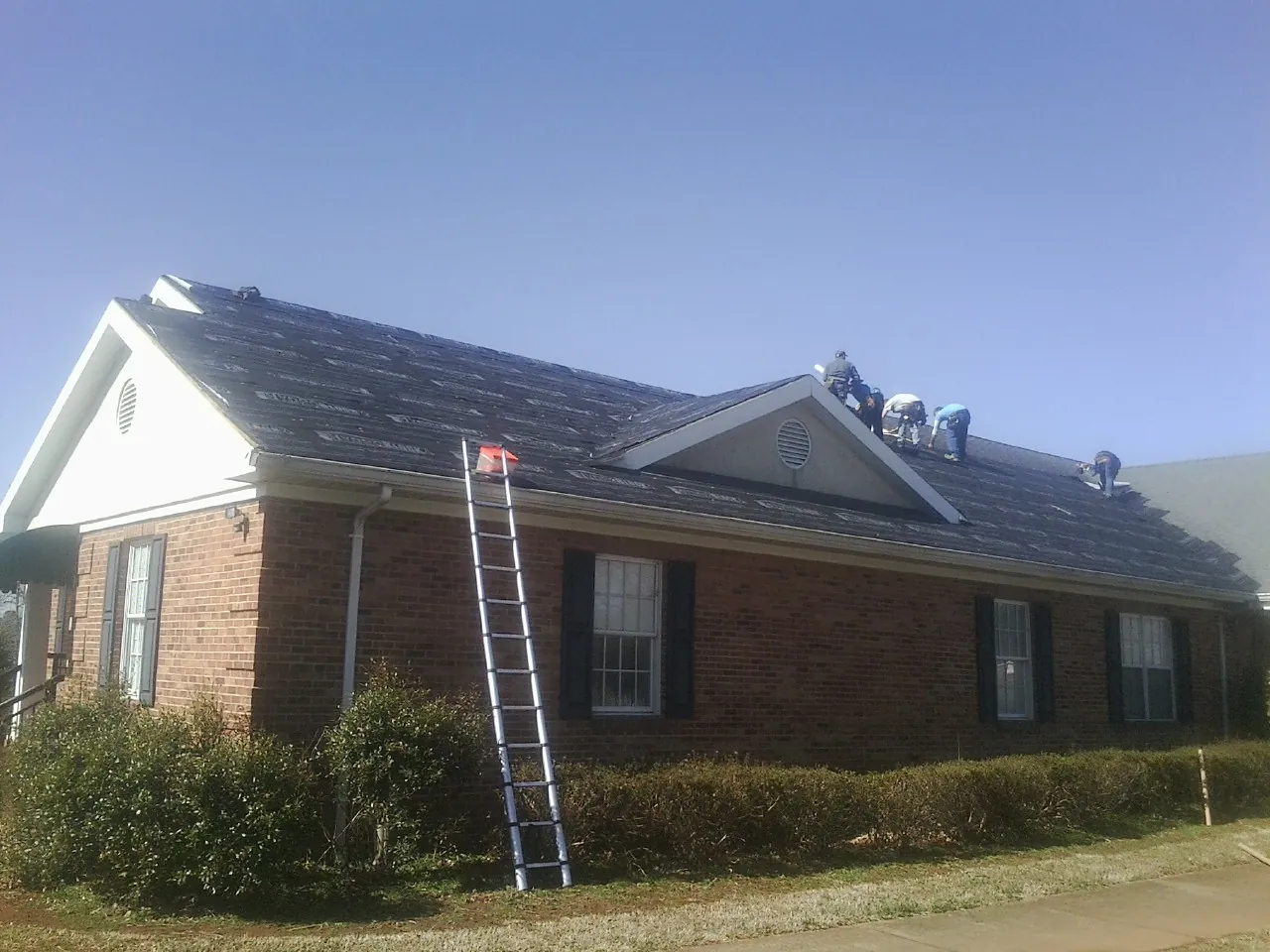 Crew tearing shingles off a brick ranch roof with a ladder set — Metal Roof Repair work by our crew in Swanville