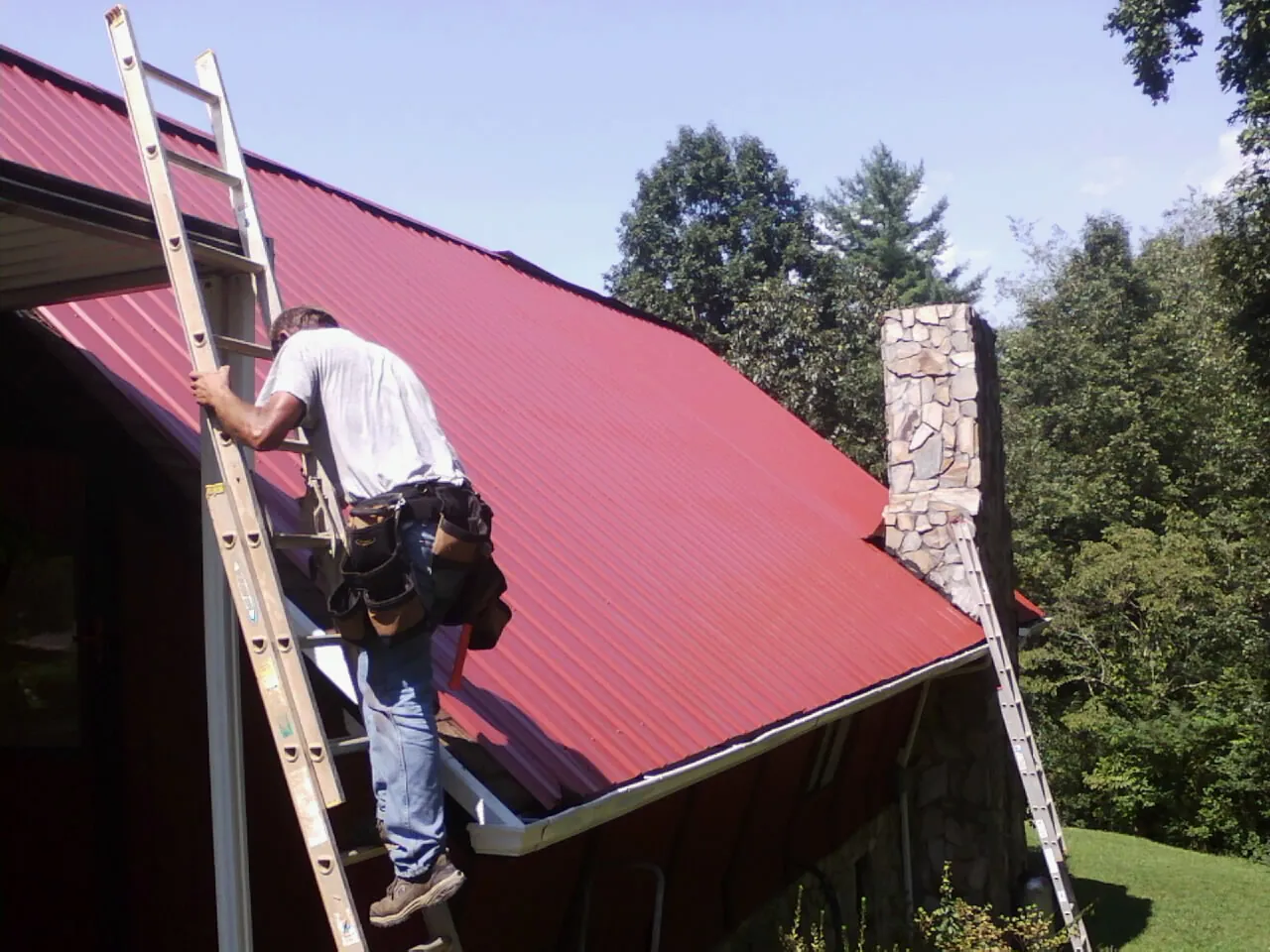 Roofer on a ladder beside a red metal roof and stone chimney — Soffit & Fascia Repair by Roofing Brothers in Swanville